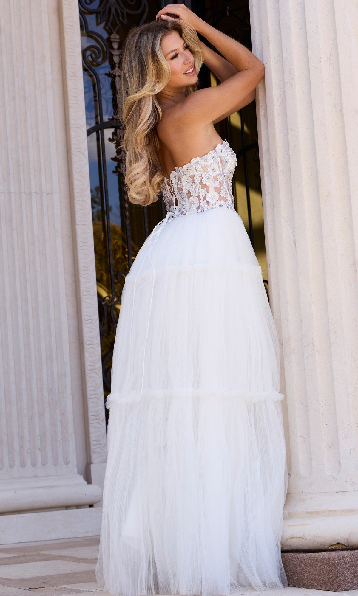 Wearing the Jovani 45866 long prom dress with a strapless corset bodice and flowing tulle A-line skirt, a woman stands by a column, smiling over her shoulder.